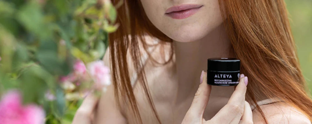 A woman with red hair surrounded by rose fields, holding a bottle of Alteya Rosa Damascena cream.