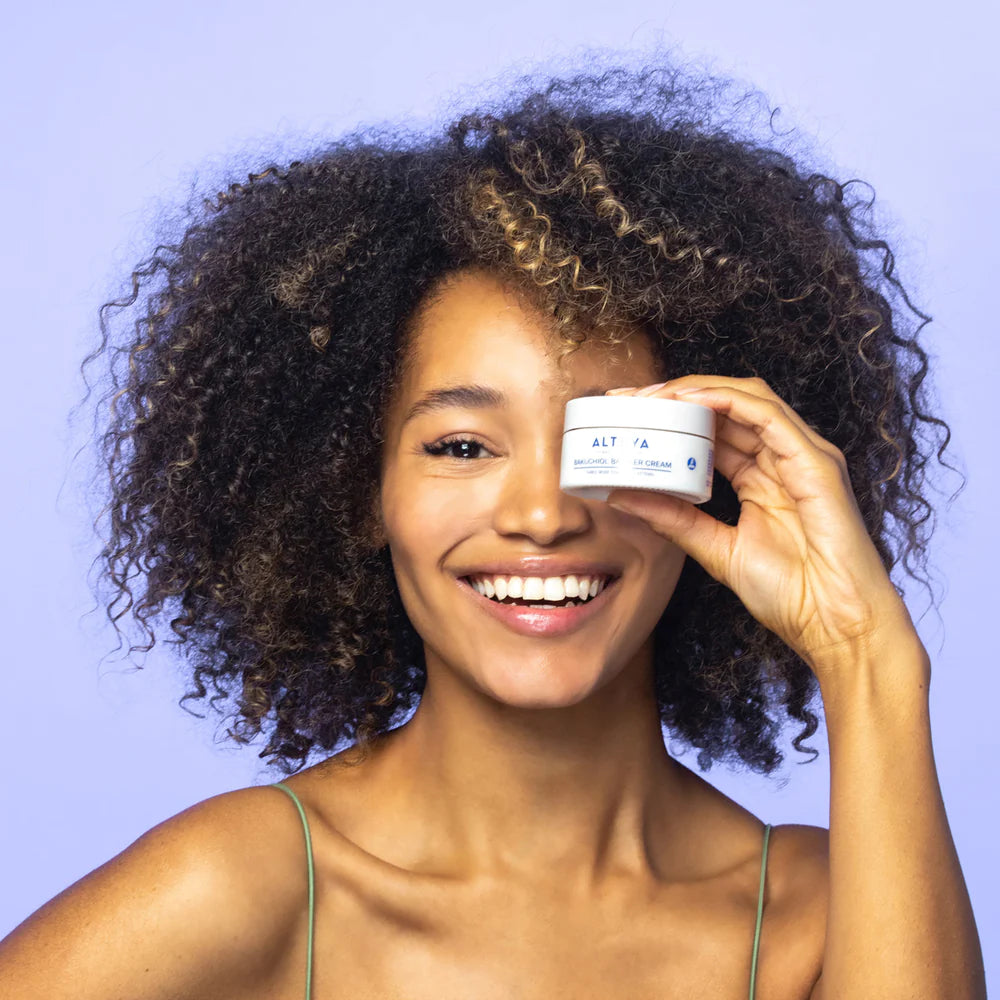 A smiling person with curly hair holds a small container labeled "ALTA" in front of their face against a light purple background, showcasing the Rose Hydrobiome Bakuchiol Barrier Cream for radiant skin.