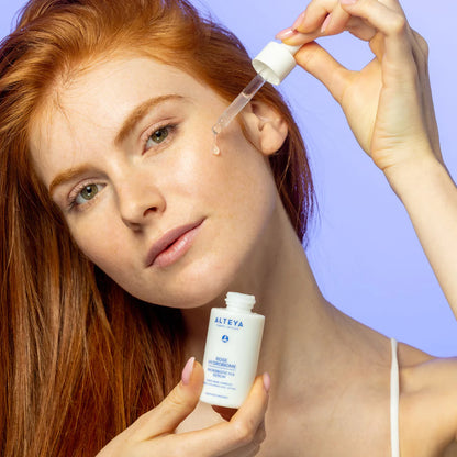 A person with red hair is holding a dropper and a bottle of Rose Hydrobiome Microbiotic H.A. Serum, dispensing a liquid onto their face.