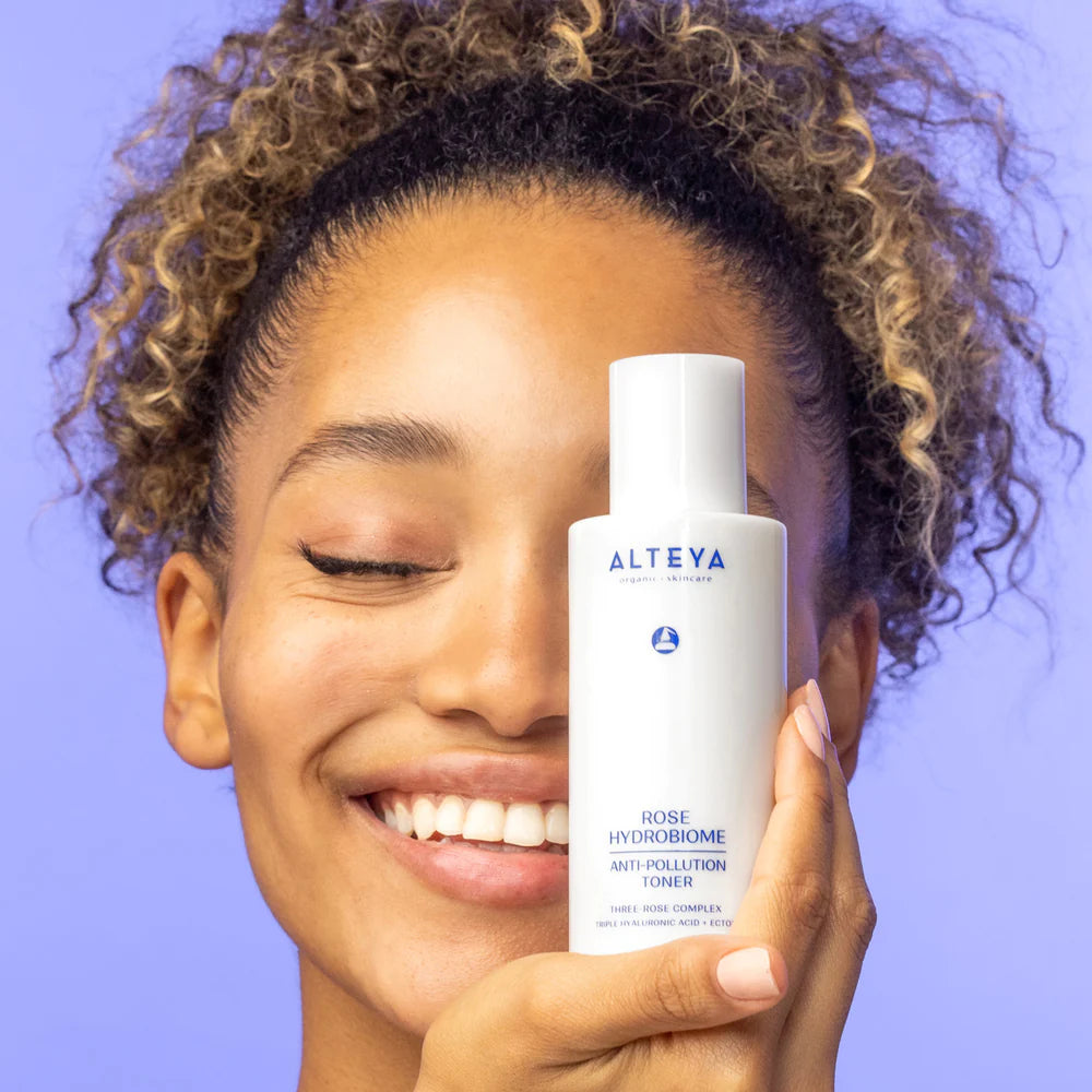 A woman smiling while holding a bottle of Rose Hydrobiome Anti-Pollution Toner with her eyes closed.