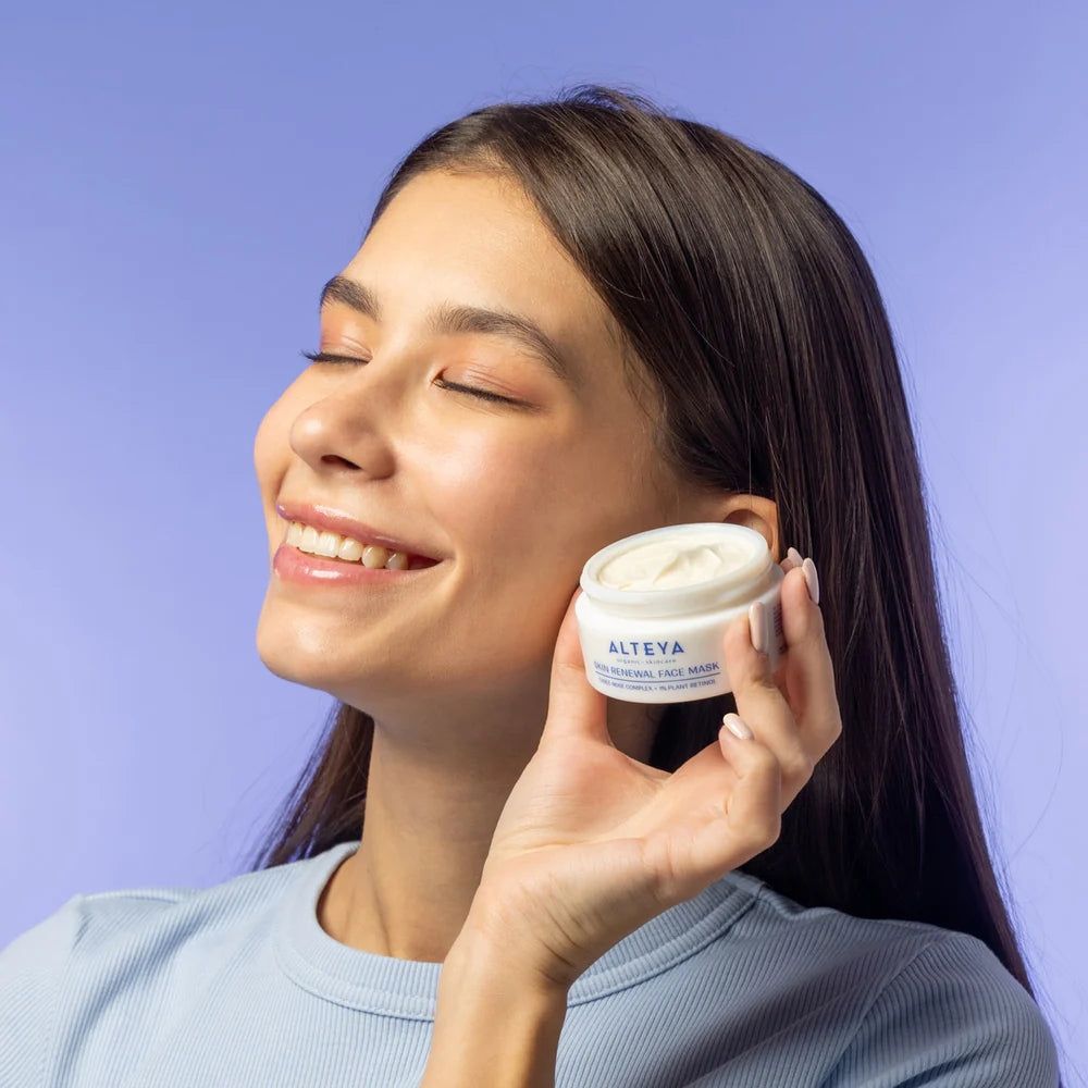 A woman smiles with closed eyes, holding a jar of Rose Hydrobiome Skin Renewal Face Mask close to her face. She is wearing a light blue top against a pastel purple background, basking in the natural glow from the mask&