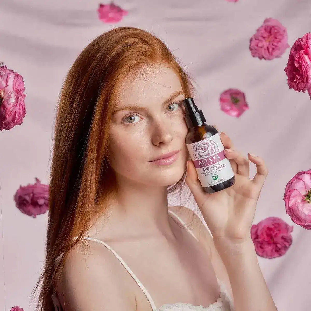 A woman with long red hair holds a bottle of Alteya Organics rose water; pink roses are scattered on a pale pink background.