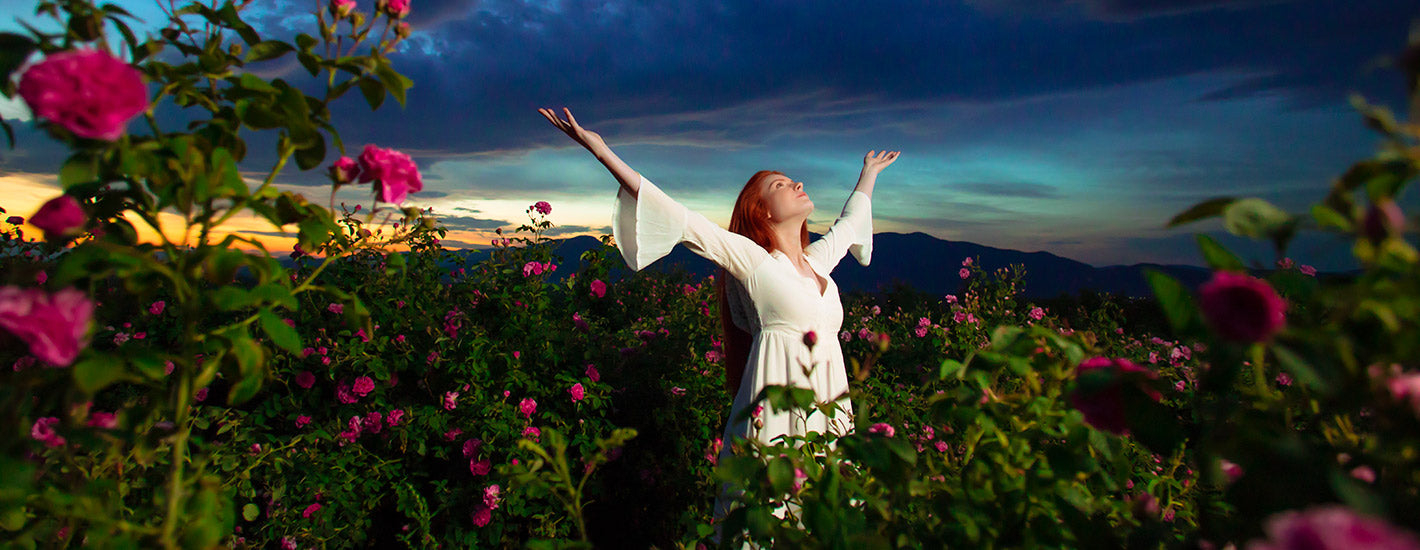 A woman standing in a field of rose fields with her arms outstretched, surrounded by rosa damascena and alteya roses.
