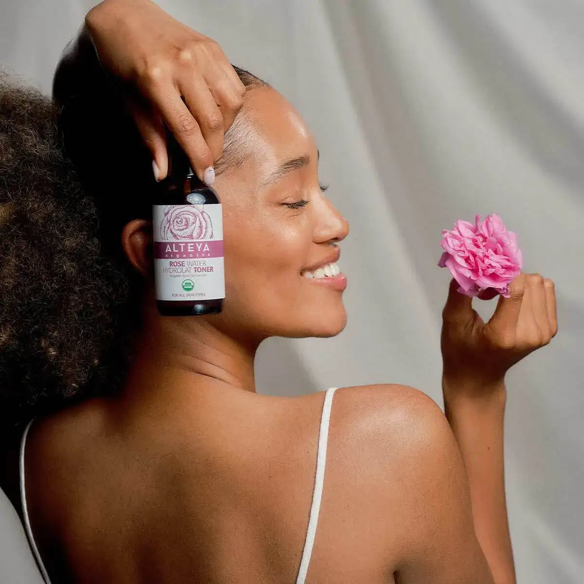 A person smiles warmly, holding a pink rose and a bottle labeled &quot;Bulgarian Organic Rose Water – Glass Spray,&quot; expertly crafted from organic rose water sourced from the renowned Bulgarian Rose Valley, set against a plain background.